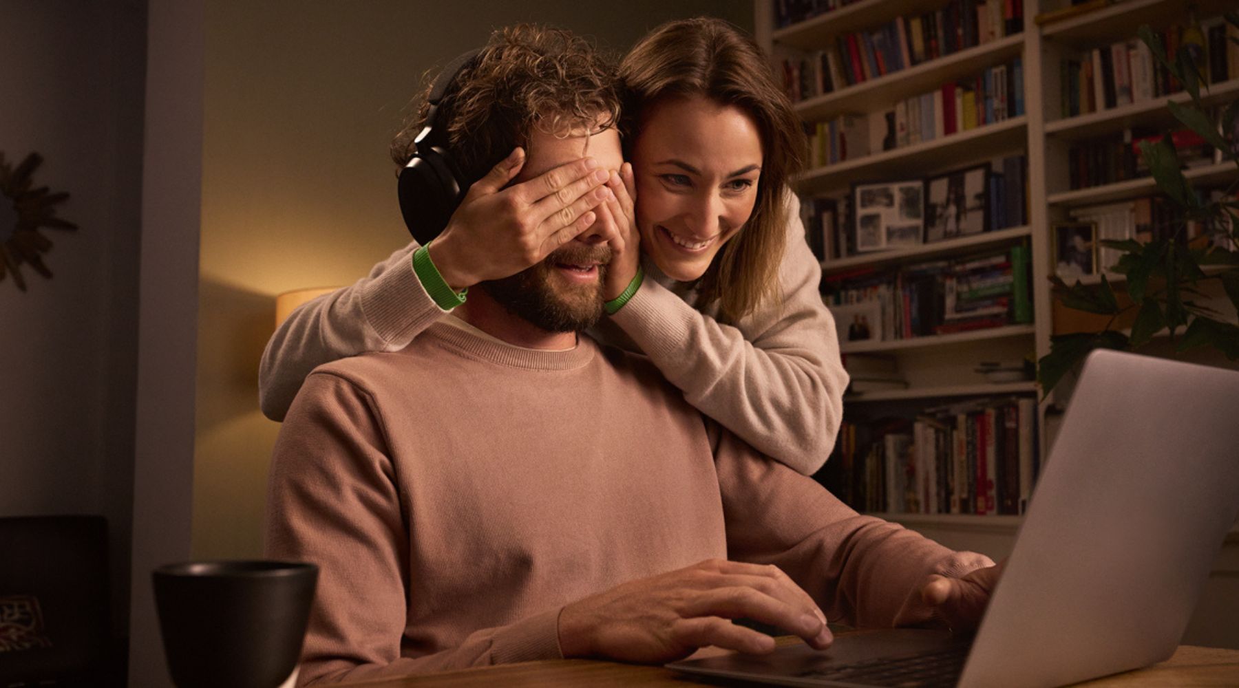 Frau hält einem Mann mit Kopfhörern vor einem Laptop spielerisch die Augen zu.